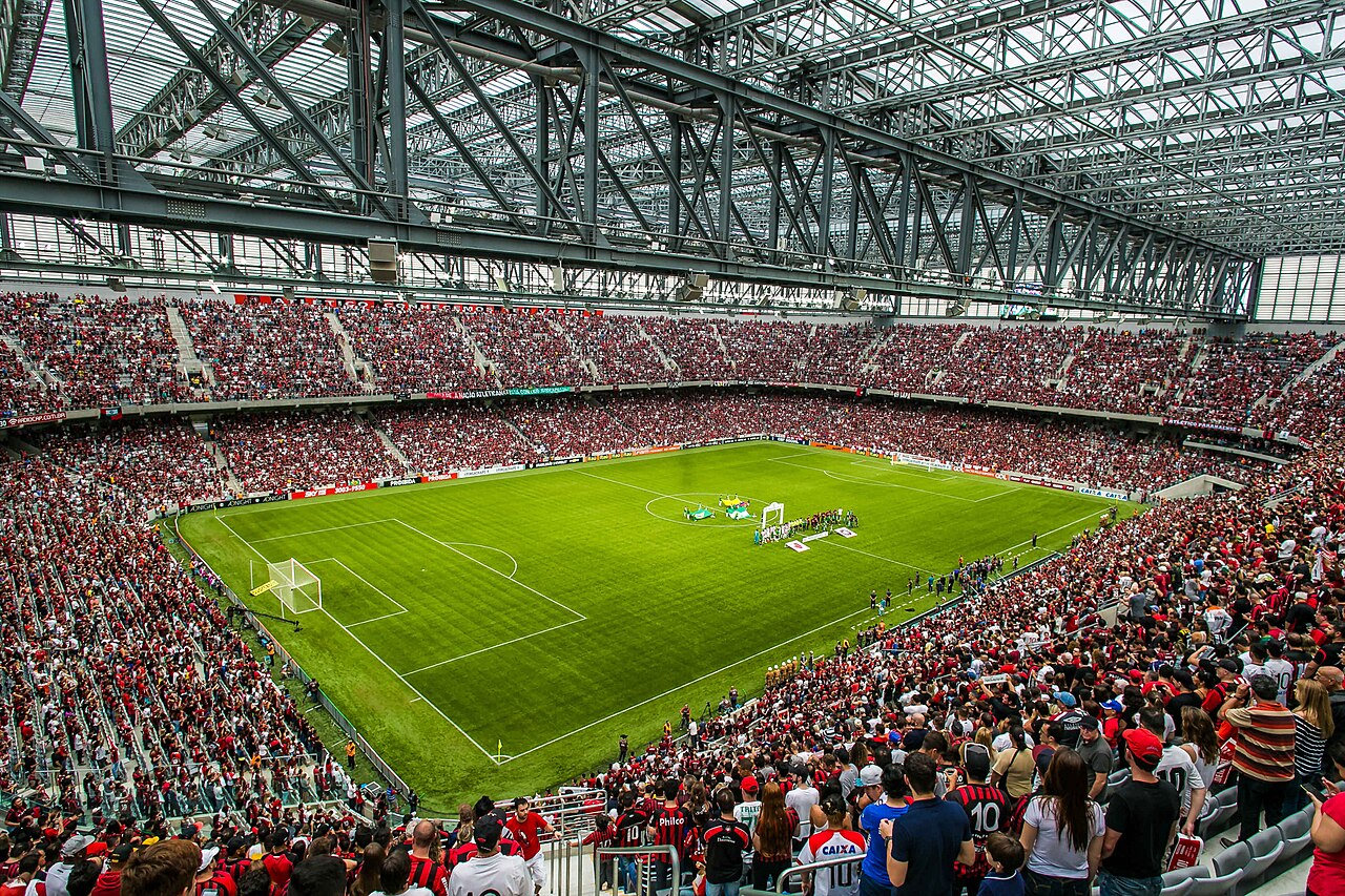 Imagem ilustrativa: Ligga Arena, estádio do Club Athletico Paranaense, em Curitiba, com arquibancadas lotadas e o gramado antes de uma partida.