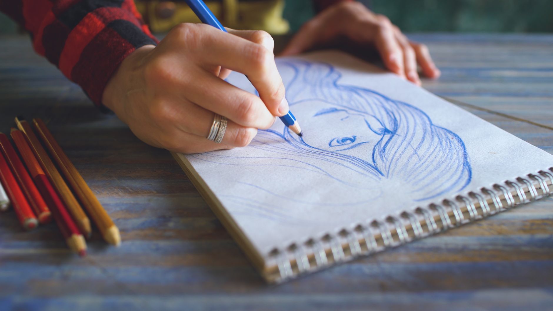 Imagem ilustrativa: Mão desenhando um rosto a lápis em caderno espiral, com lápis de cor sobre a mesa