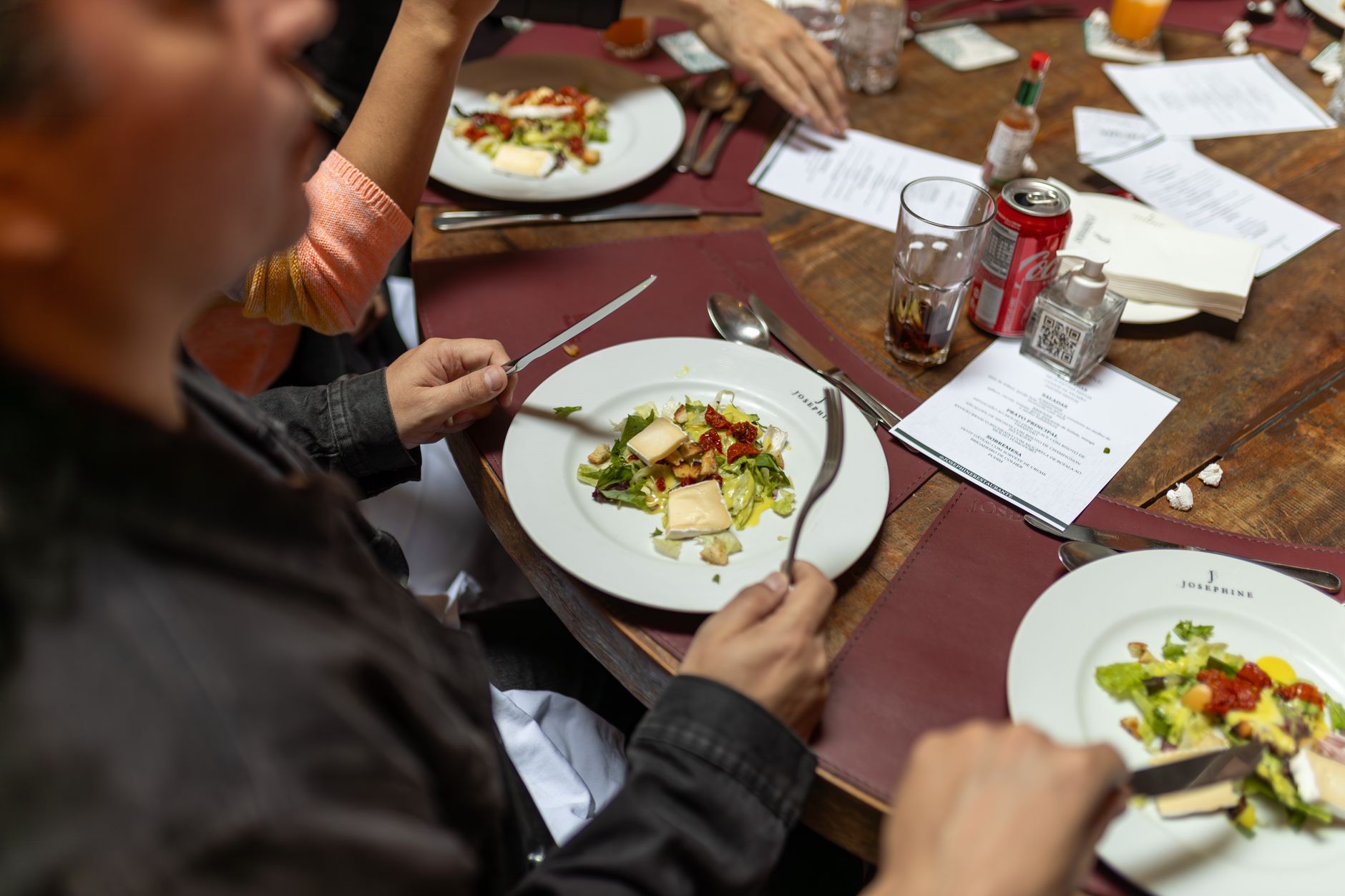Imagem ilustrativa: Mesa de restaurante com pratos servidos, cardápios e clientes comendo