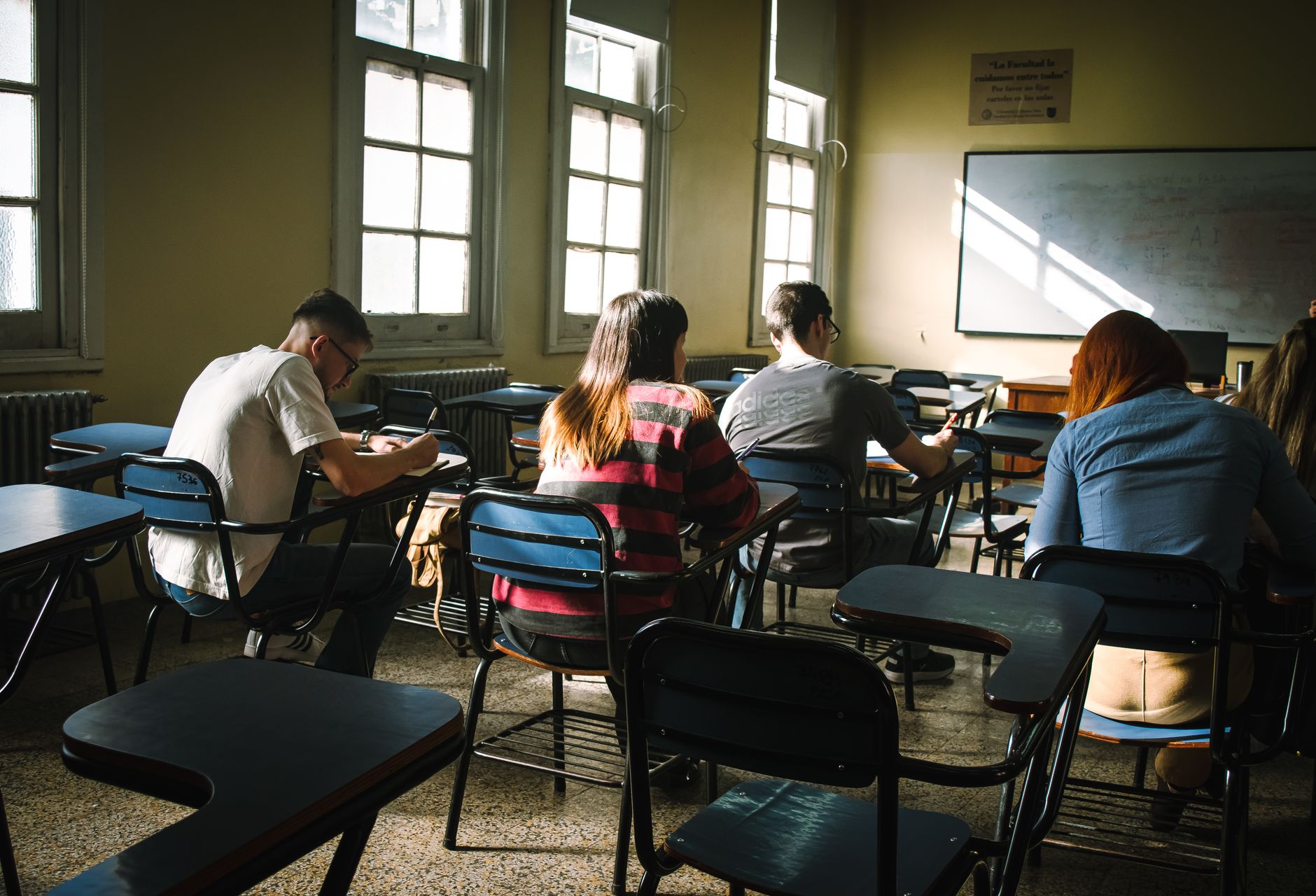Imagem ilustrativa: Um grupo de alunos concentrados na escrita em uma sala de aula ensolarada, com carteiras e janelas.