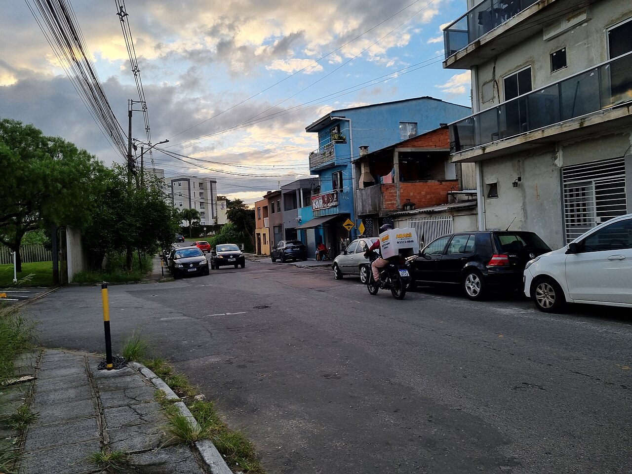 Imagem ilustrativa: Rua Rogério Pereira de Camargo, no bairro Cidade Industrial de Curitiba, com carros estacionados e uma moto em circulação.