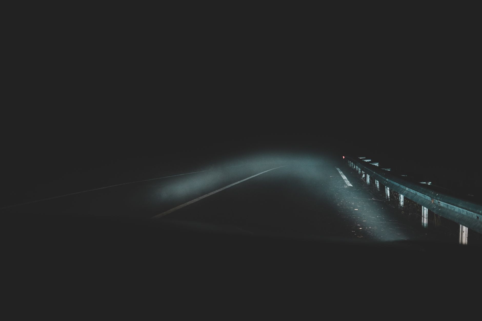 Imagem ilustrativa: Estrada noturna sombria com neblina e guard-rails visíveis, iluminada pelos faróis do carro.