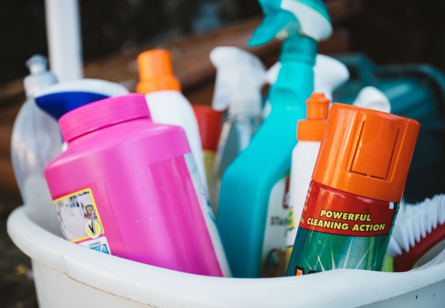 Imagem ilustrativa: Close-up de diversos produtos de limpeza em um balde ao ar livre, exibindo vários detergentes e sprays.