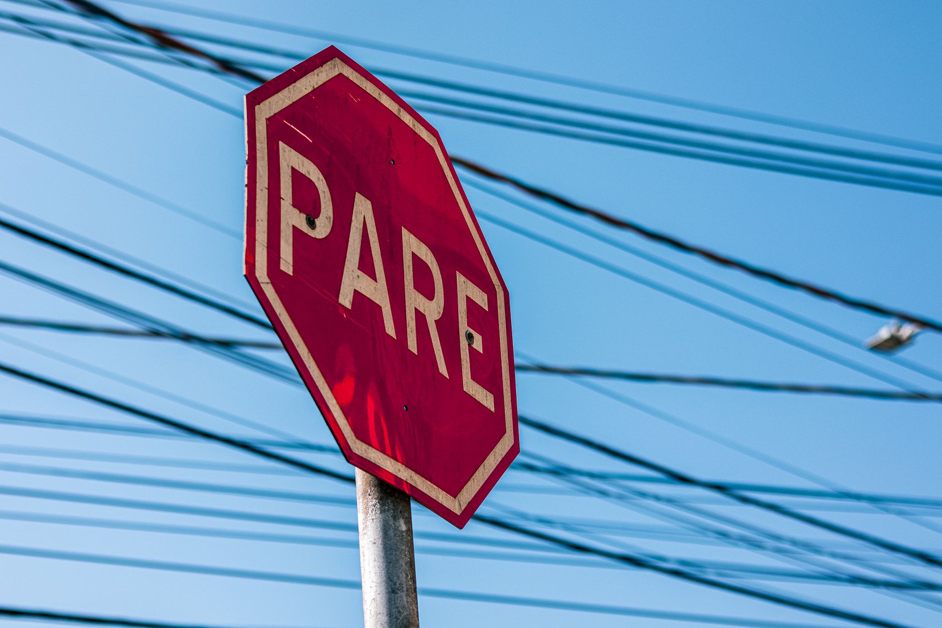 Imagem ilustrativa: Red stop sign with 'PARE' text against a clear blue sky and overhead power lines.