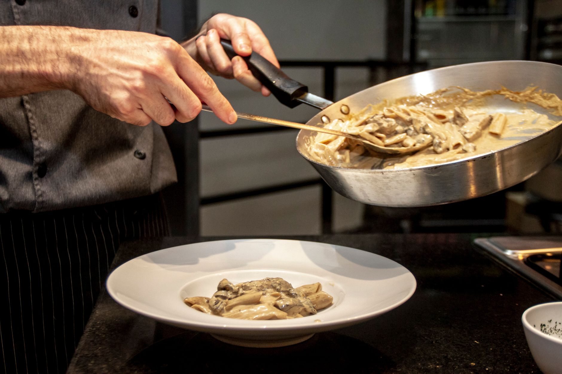 Imagem ilustrativa: Chef cuidadosamente monta um prato de massa cremosa com cogumelos em um ambiente de cozinha interna.