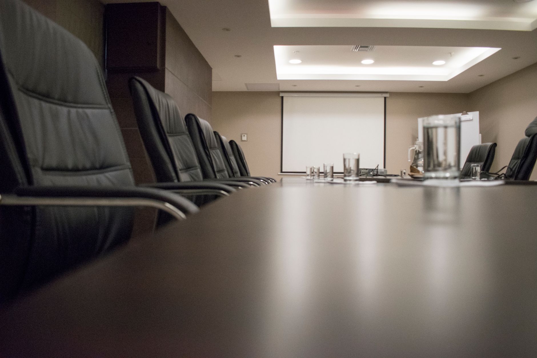 Imagem ilustrativa: Sala de reunião vazia com mesa longa, cadeiras, tela de projeção e copos sobre a mesa