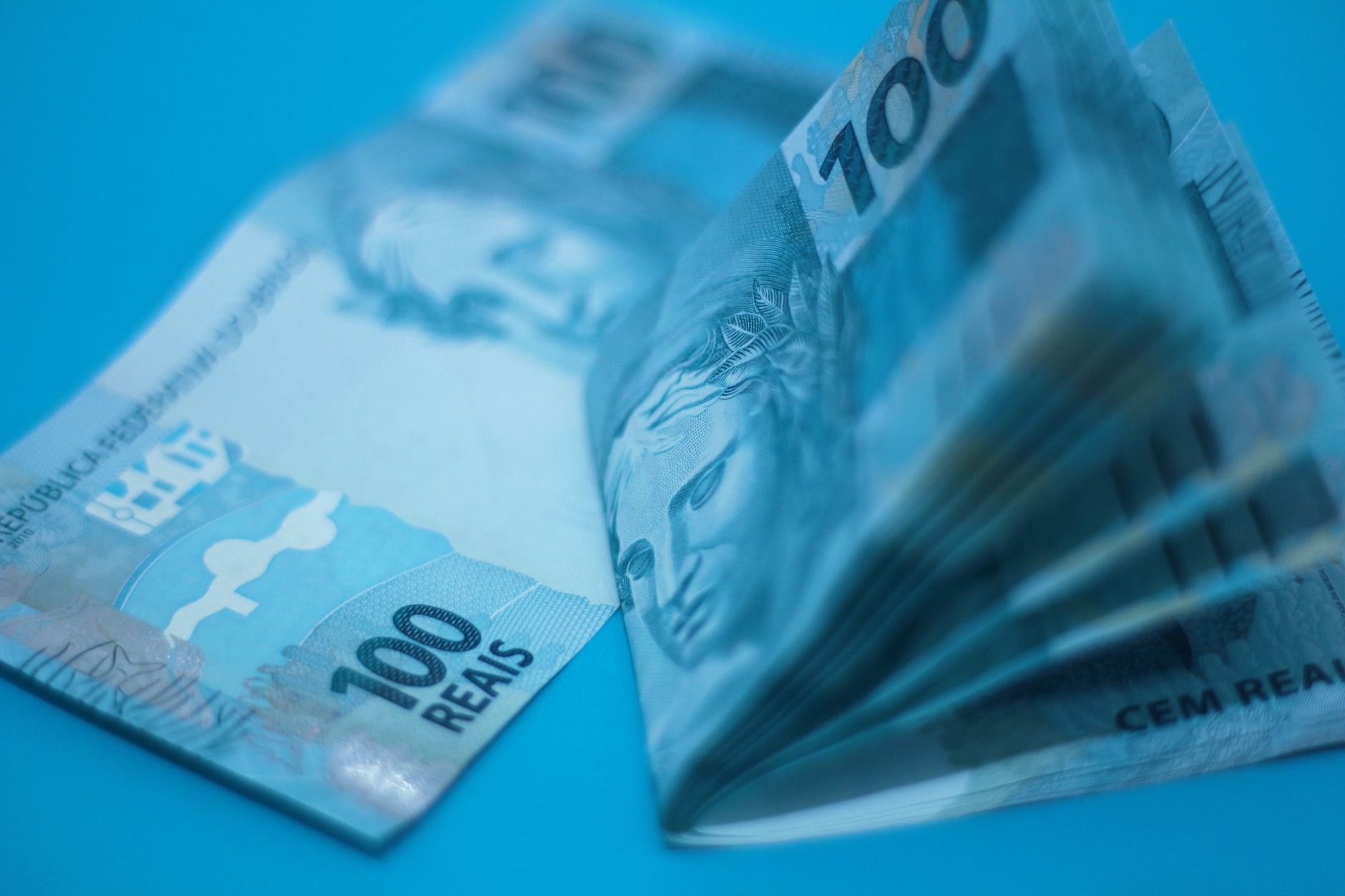 Imagem ilustrativa: Notas de cem reais sobre superfície azul, em primeiro plano