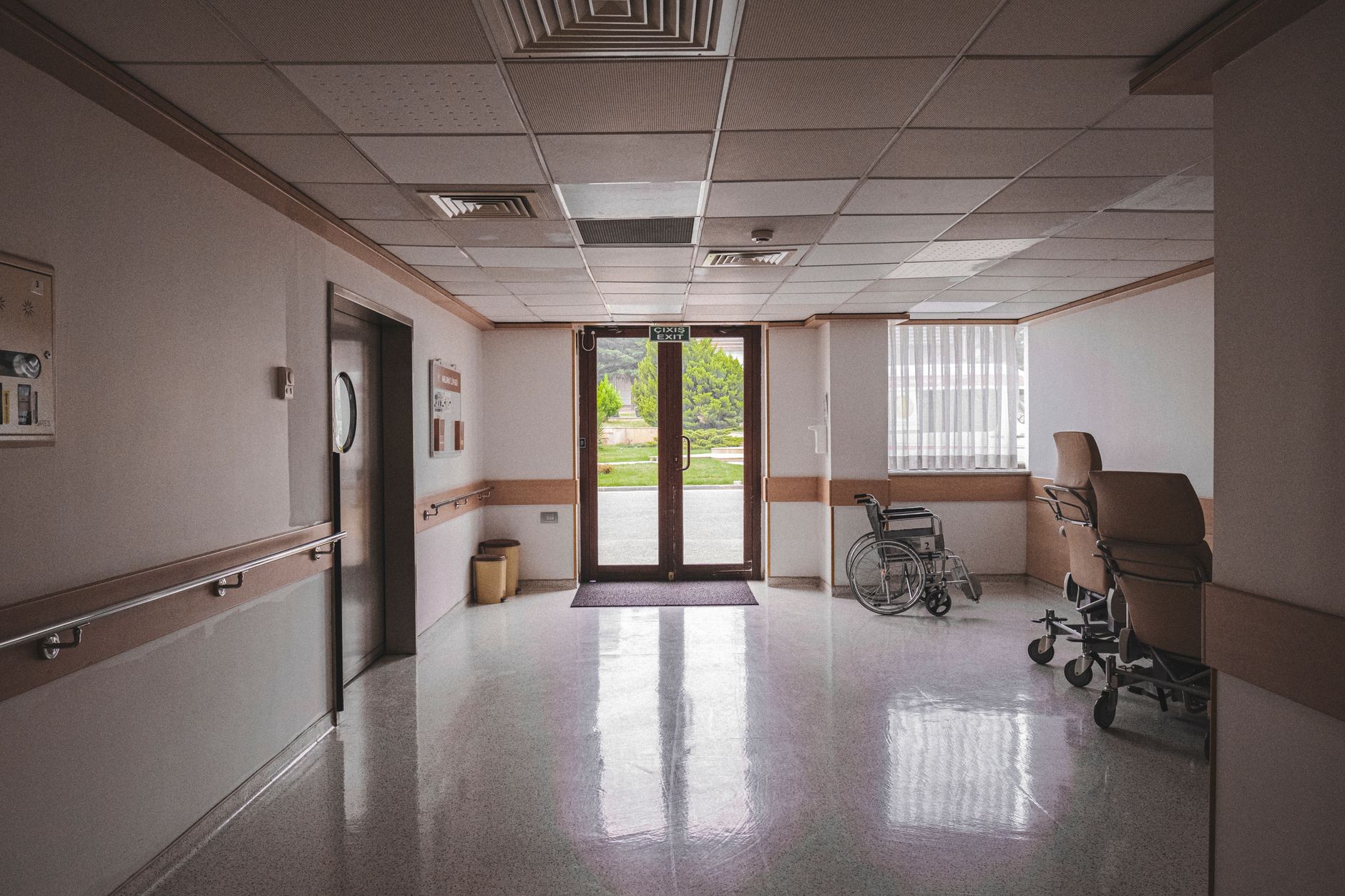 Imagem ilustrativa: Corredor de hospital com cadeira de rodas, portas de vidro refletivas e vista externa.